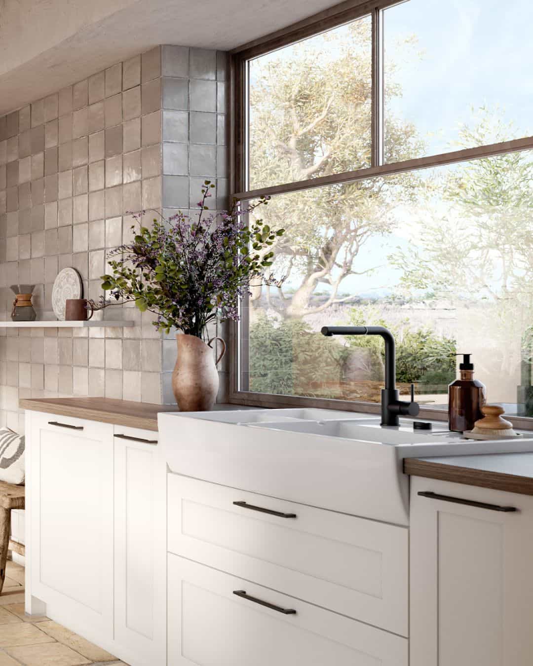 Cuisine classique blanche avec façades à cadres élégantes, poignées en métal, plan de travail effet bois naturel, évier timbre d’office, carrelage beige mural et grande fenêtre
