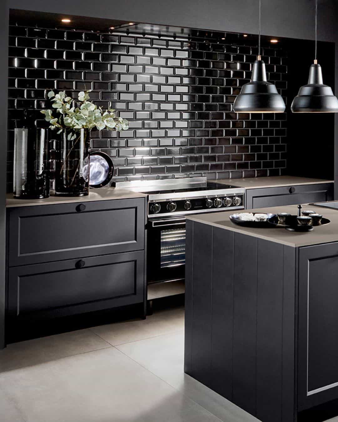 Cuisine classique noire avec îlot central, façades à cadres, cuisinière grand format au style professionnel, luminaires suspendus vintage en métal noir et crédence en carrelage noir brillant. IDEO cuisines sur mesure