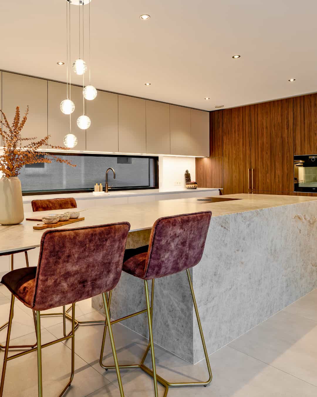 Cuisine design haut de gamme avec grand îlot central en céramique effet marbre, façades en bois avec four encastré et espace repas avec tabourets en velours. IDEO Cuisine équipée sur mesure